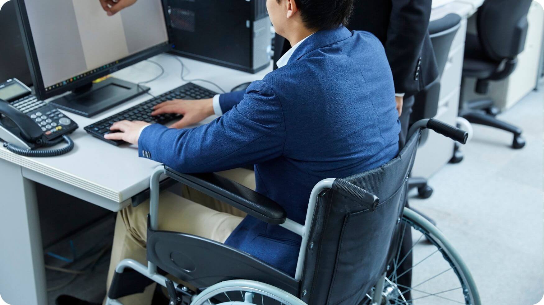A man is sat in a wheelchair working on a computer at an office desk.
