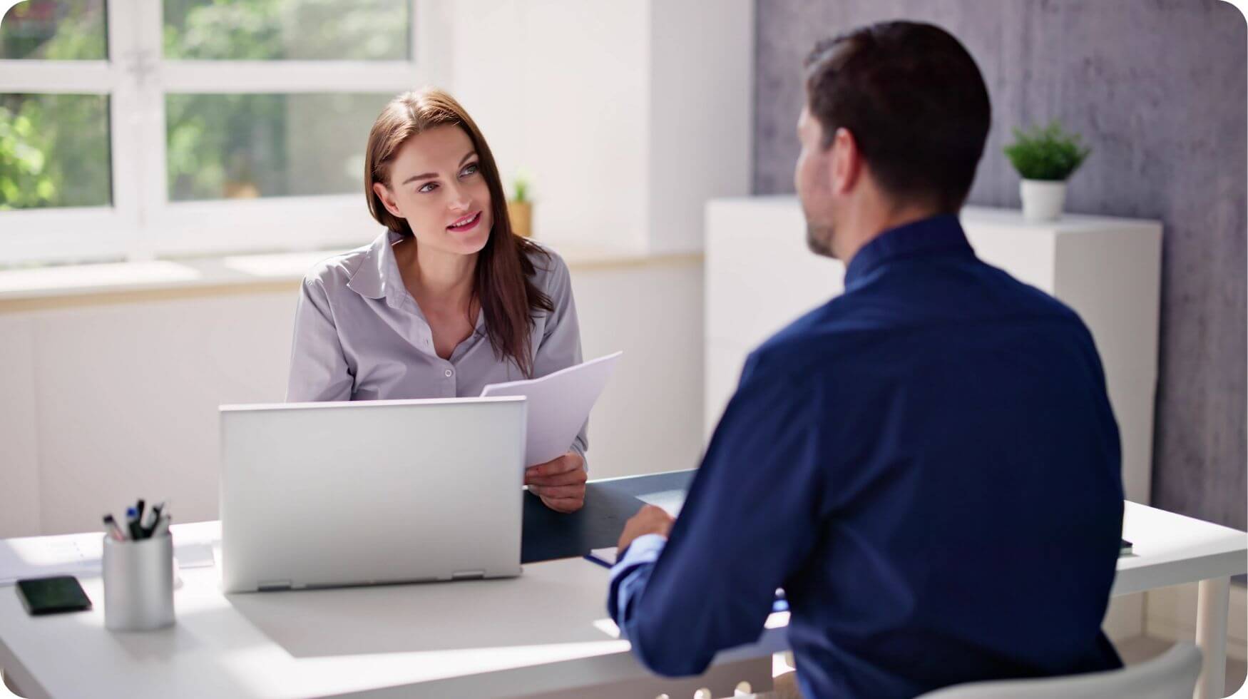 A woman is sat behind a desk holding a document and interviewing a man.
