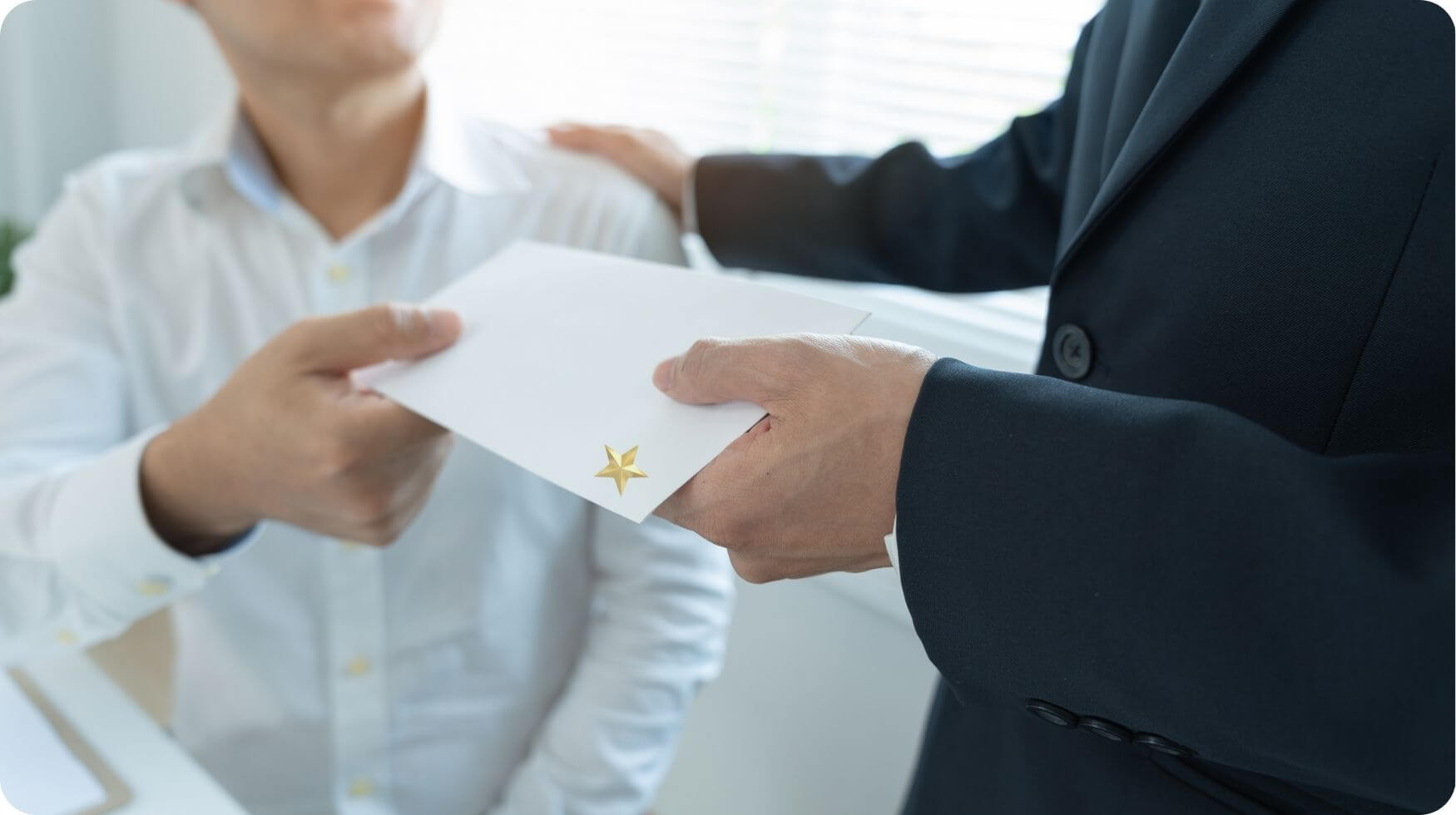 A close up of a man's hands holding a white envelope which is being handed to a man sat down.