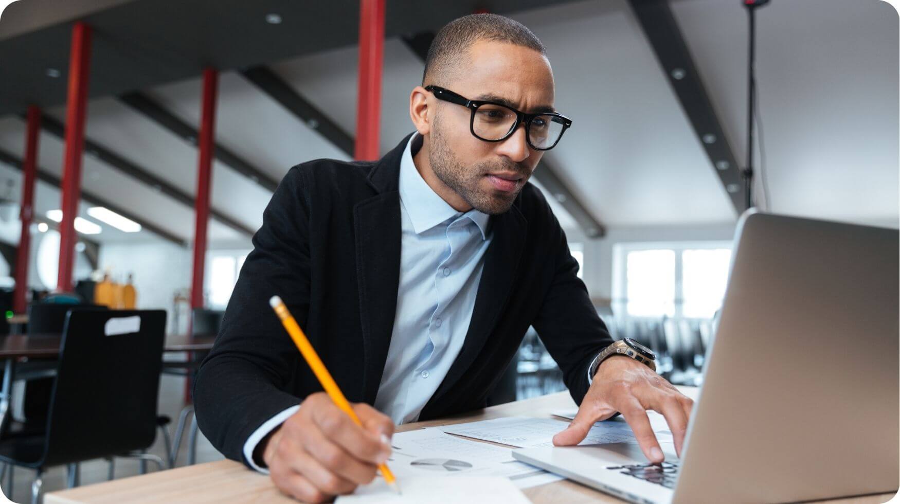 A man wearing glasses is looking intently at a computer screen whilst holding a yellow pencil and making notes.