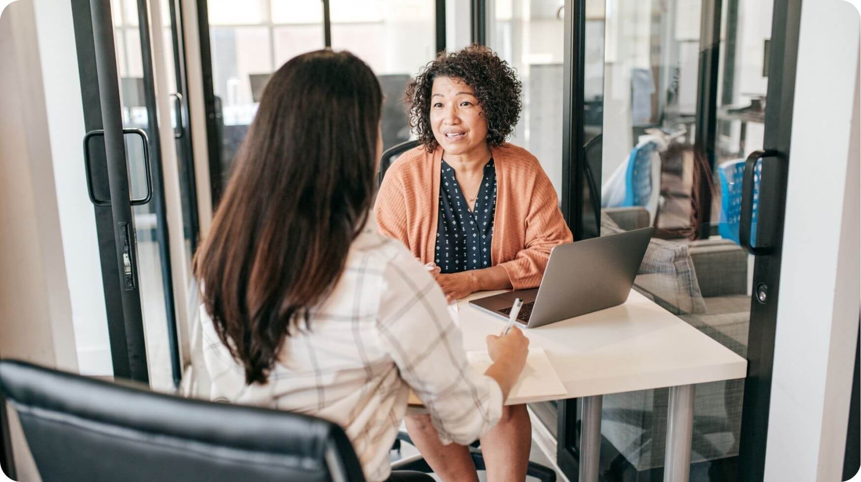 Two women are sat facing each other at a desk in a job interview style setting.
