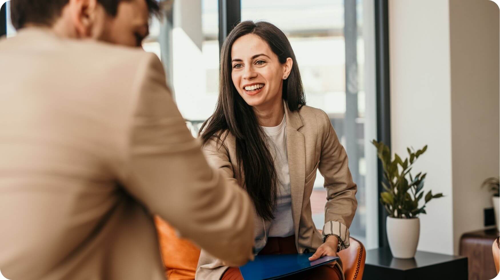 A smiling woman is holding a blue folder and shaking hands with a man who is sat with his back to the camera.