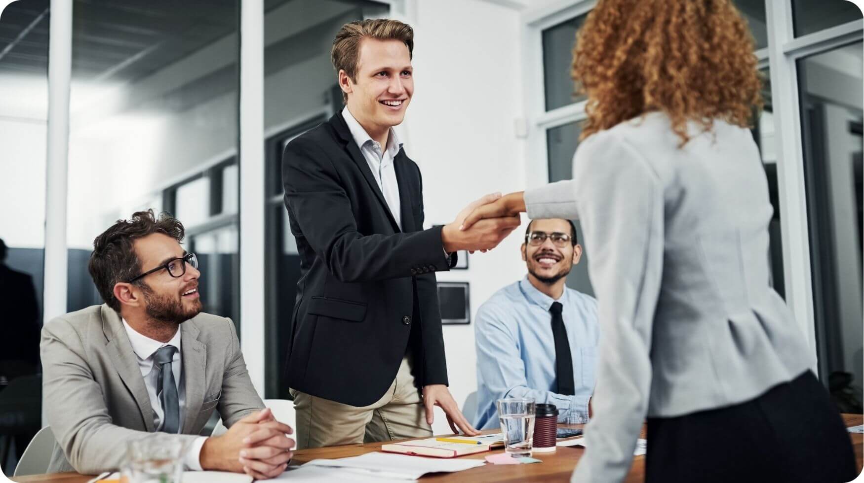 A man is standing up smiling and shaking hands with a woman. There are two men sat down opposite the woman.