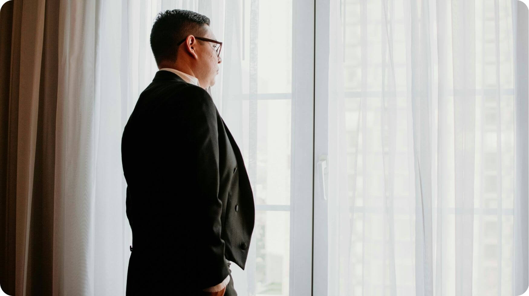 A man wearing a suit is stood facing a window which is covered with net curtains.