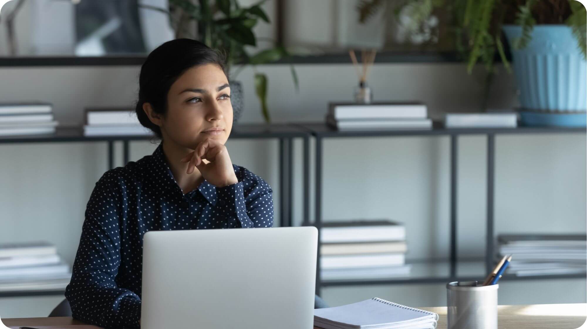 A woman is sat at a desk with a laptop in front of her. She has her chin resting on her hand and is looking thoughtful.