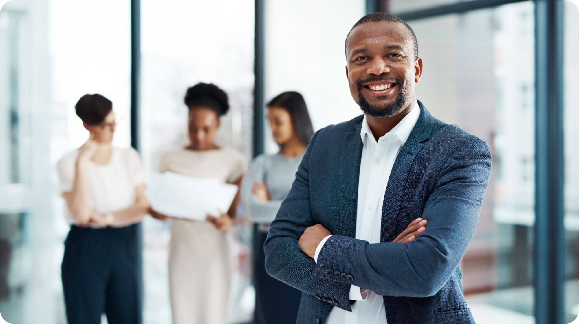 There is a smiling man in the foreground with folded arms and three women in the background who are reviewing a document.